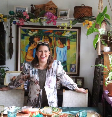 JoySauce tour: Giselle Tongi | biracial Asian/Caucasian woman with medium length brown hair smiling stands with hands on top of dining chairs amidst a colorful room with plants, paintings, and decor