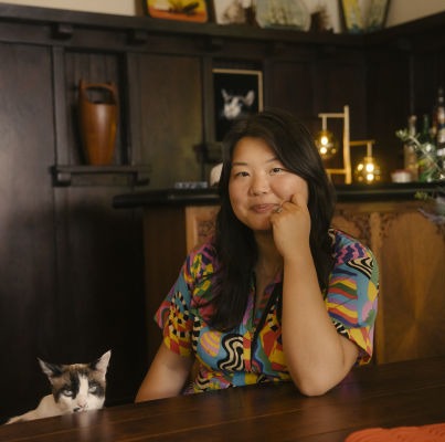 Jessica Gao | Asian American woman with long hair in pattered shirt next to gray and white cat in dark wood dining room
